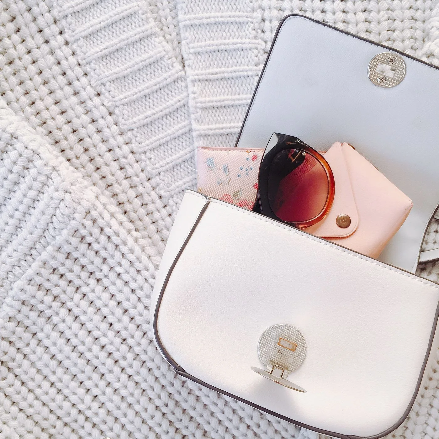 White handbag, sunglasses, and a case arranged on light-colored knitwear.