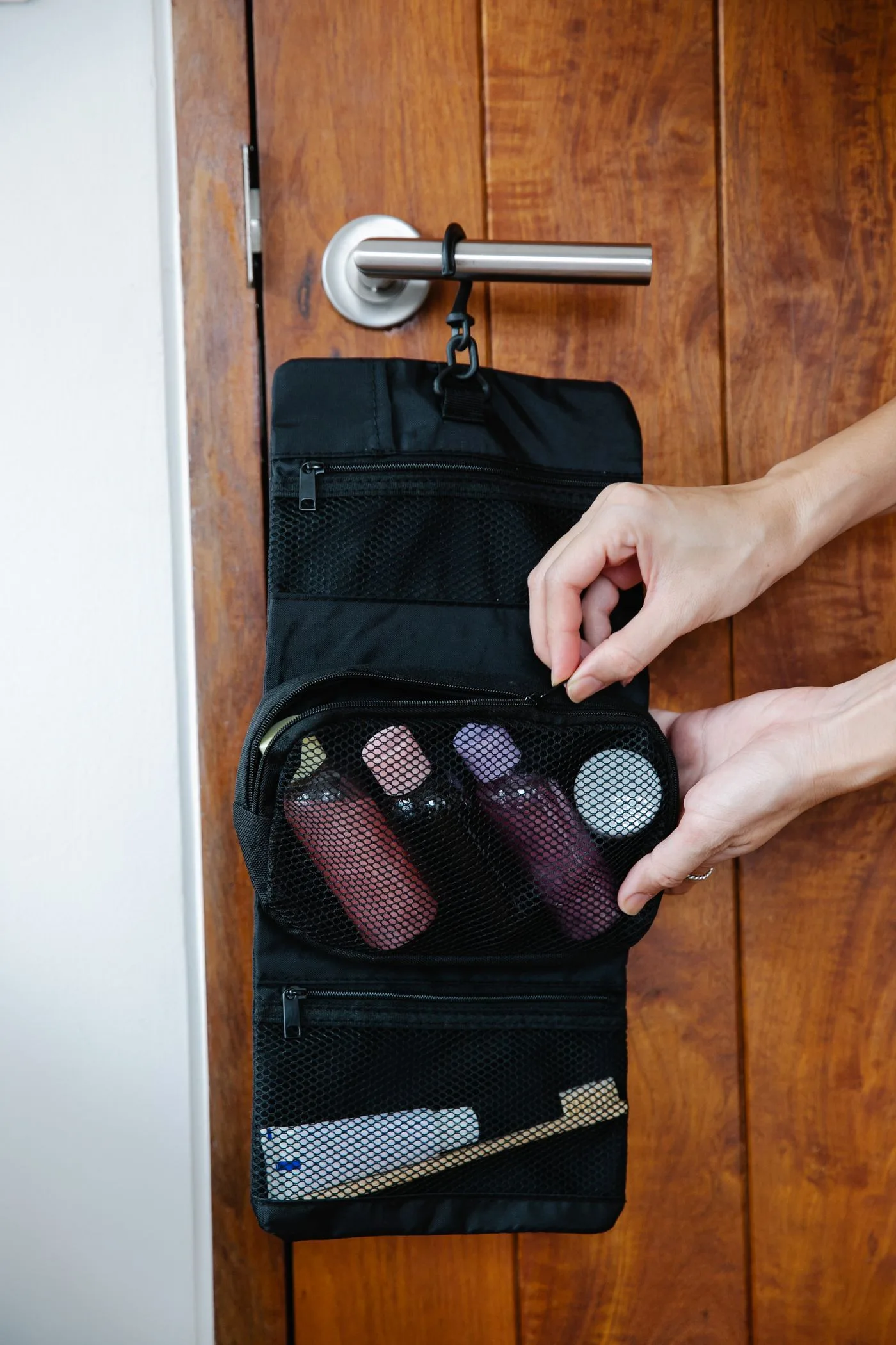 A travel toiletry bag containing cosmetic products hangs from a wooden door handle.