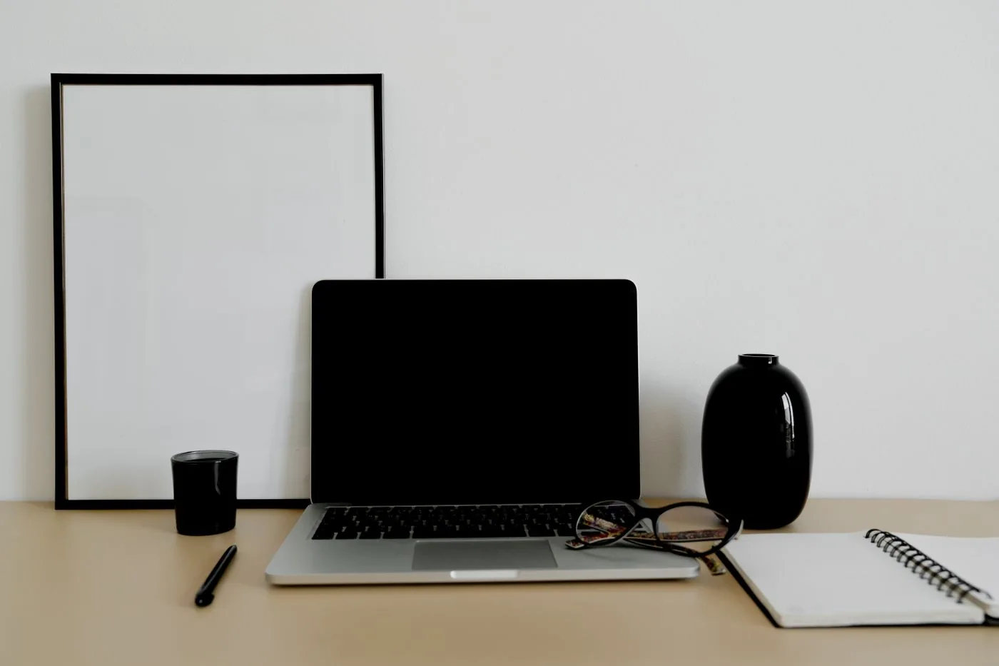 A laptop, notebook, glasses and pen on a desk beneath an empty frame