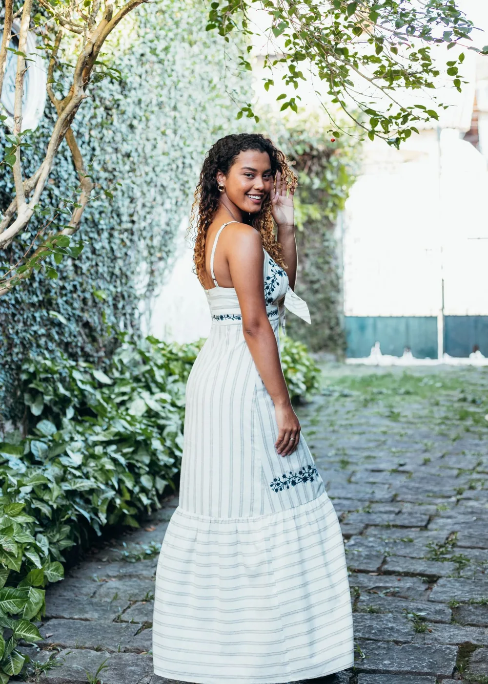 A woman in a light dress standing beside green foliage.