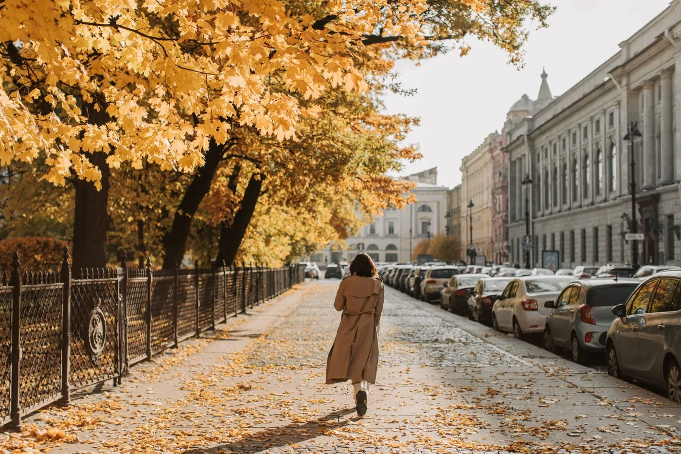 A woman in a long coat walking beside an autumn tree-lined street.