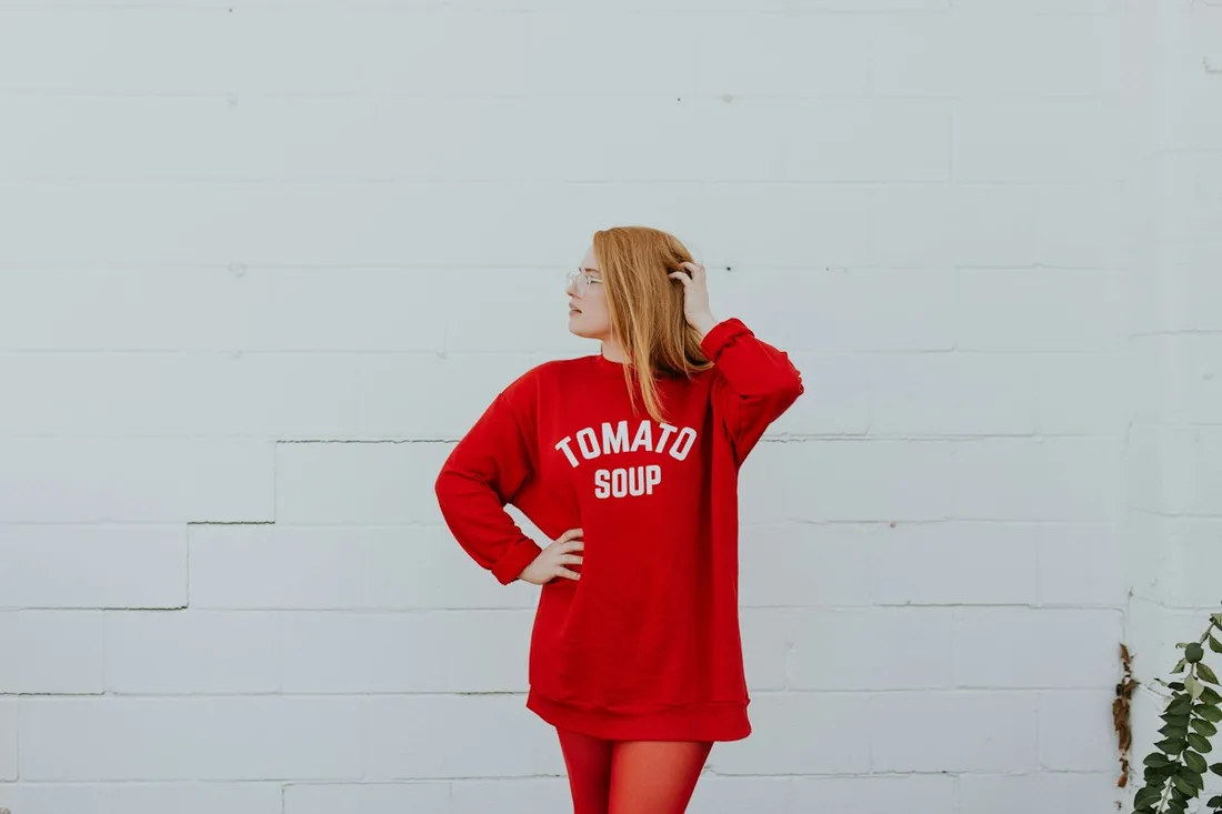 A woman wearing a red oversized sweatshirt against a pale wall.